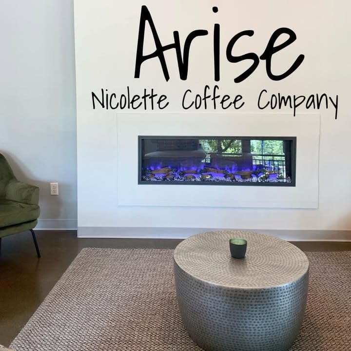 The Arise Nicolette Coffee Company name in black lettering above the long fireplace, a hammered-silver drum table in front.