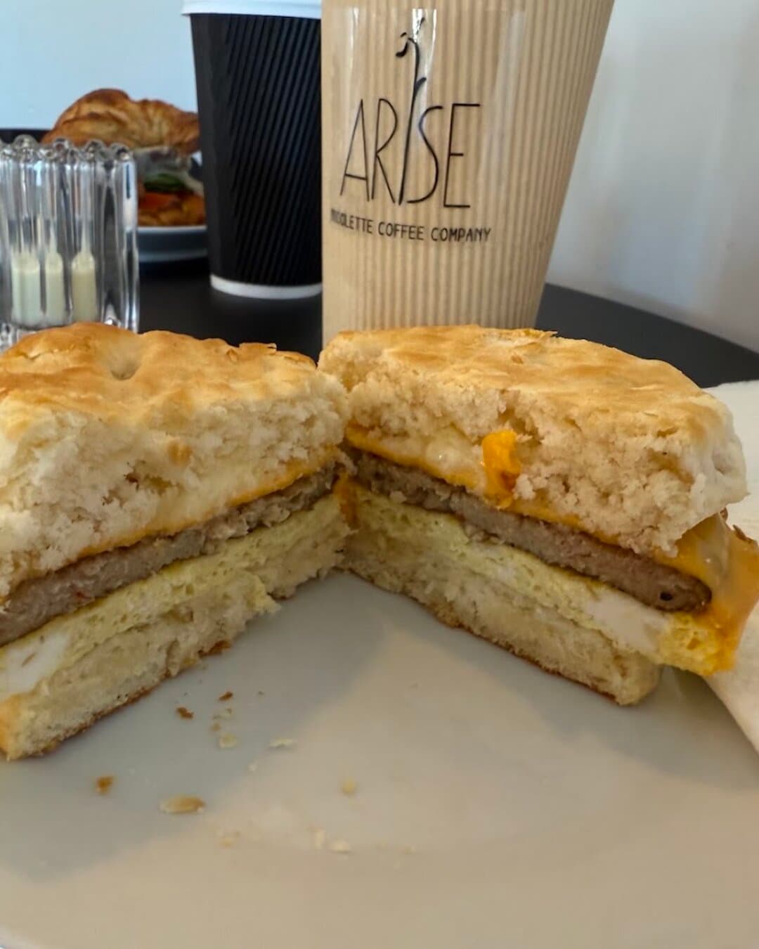 A sausage, egg and cheese breakfast sandwich on a biscuit, cut in half, an Arise cup behind it.