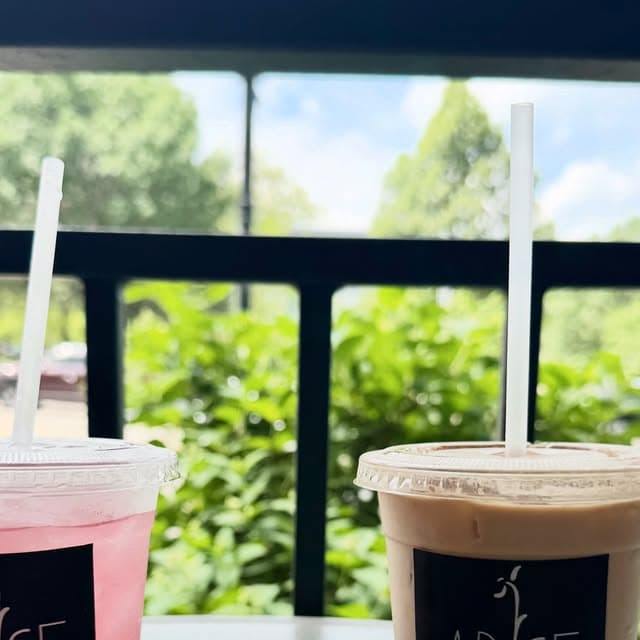 A pink refresher and an iced latte in Arise cups on the patio rail, trees behind.